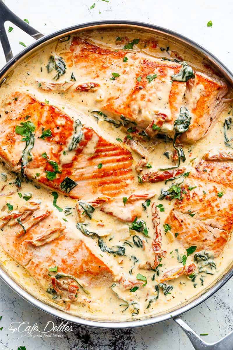 4 filetes de salmón (O TRUCHA) dorados en una sartén, en una deliciosa salsa cremosa toscana con tomates secos y espinacas marchitas, decorados con perejil o albahaca picados. | https://cafedelites.com